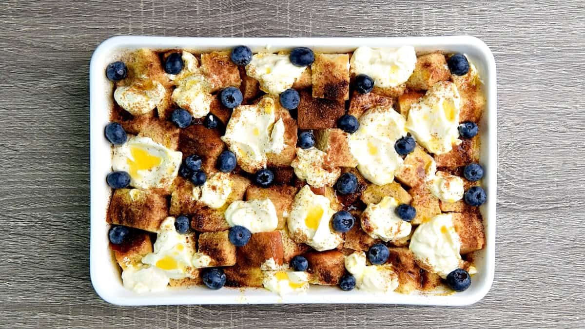 Casserole topped with cream cheese mixture, melted butter, cinnamon sugar and blueberries and ready to be baked in the oven.