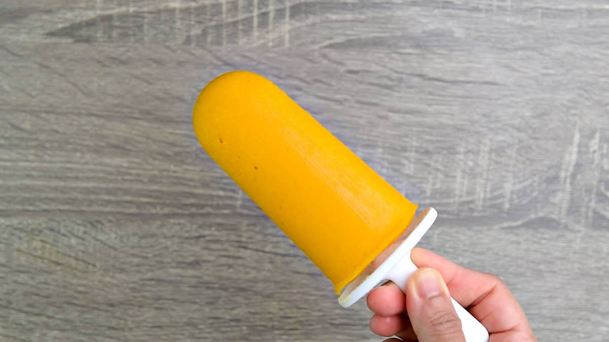 A hand holding a mango fruit popsicle fully frozen and ready to be served.
