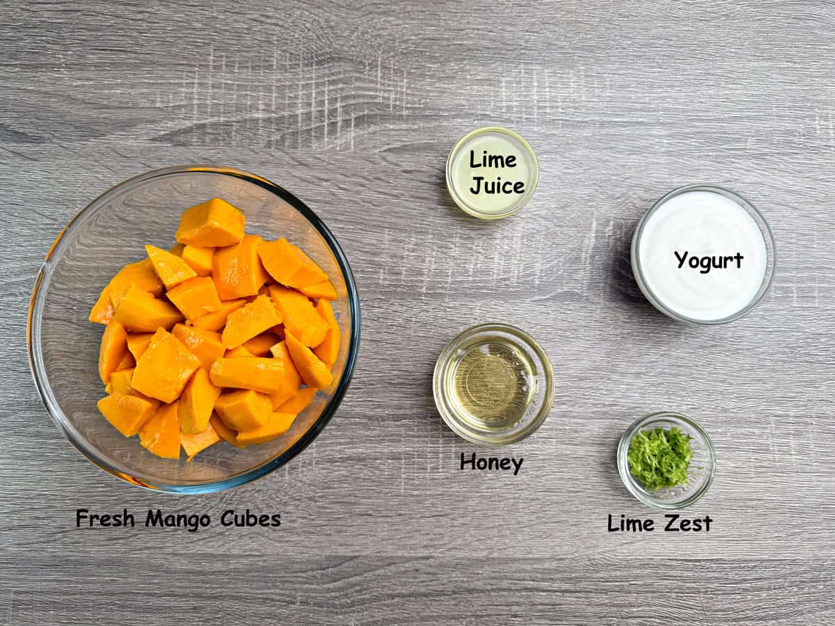 Ingredients for popsicles in bowls that include mango chunks, lime juice, yogurt, honey, lime zest, and lime.