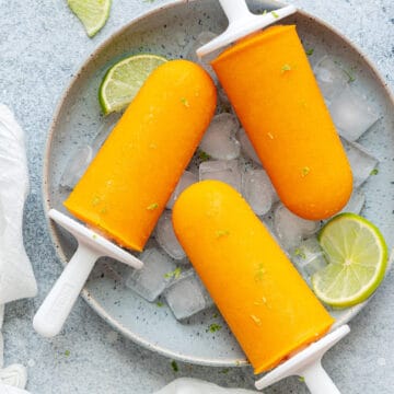 Top-down view of Mango popsicles in a bowl on ice with limes and lime zest shavings on the popsicles.