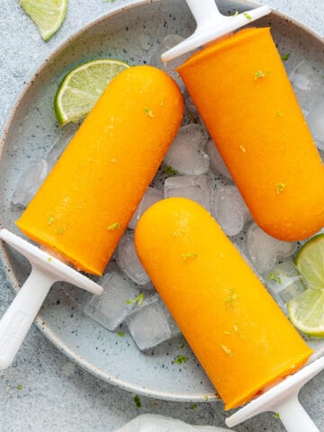 Top-down view of Mango popsicles in a bowl on ice with limes and lime zest shavings on the popsicles.