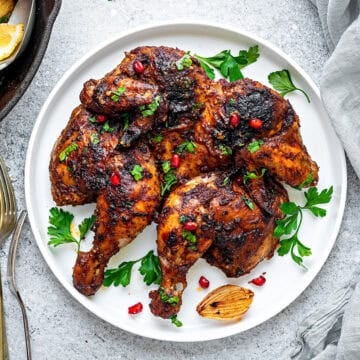 Middle Eastern flavored spatchcock chicken served on white earthen plate with roasted veggies on side in a cast iron pan.