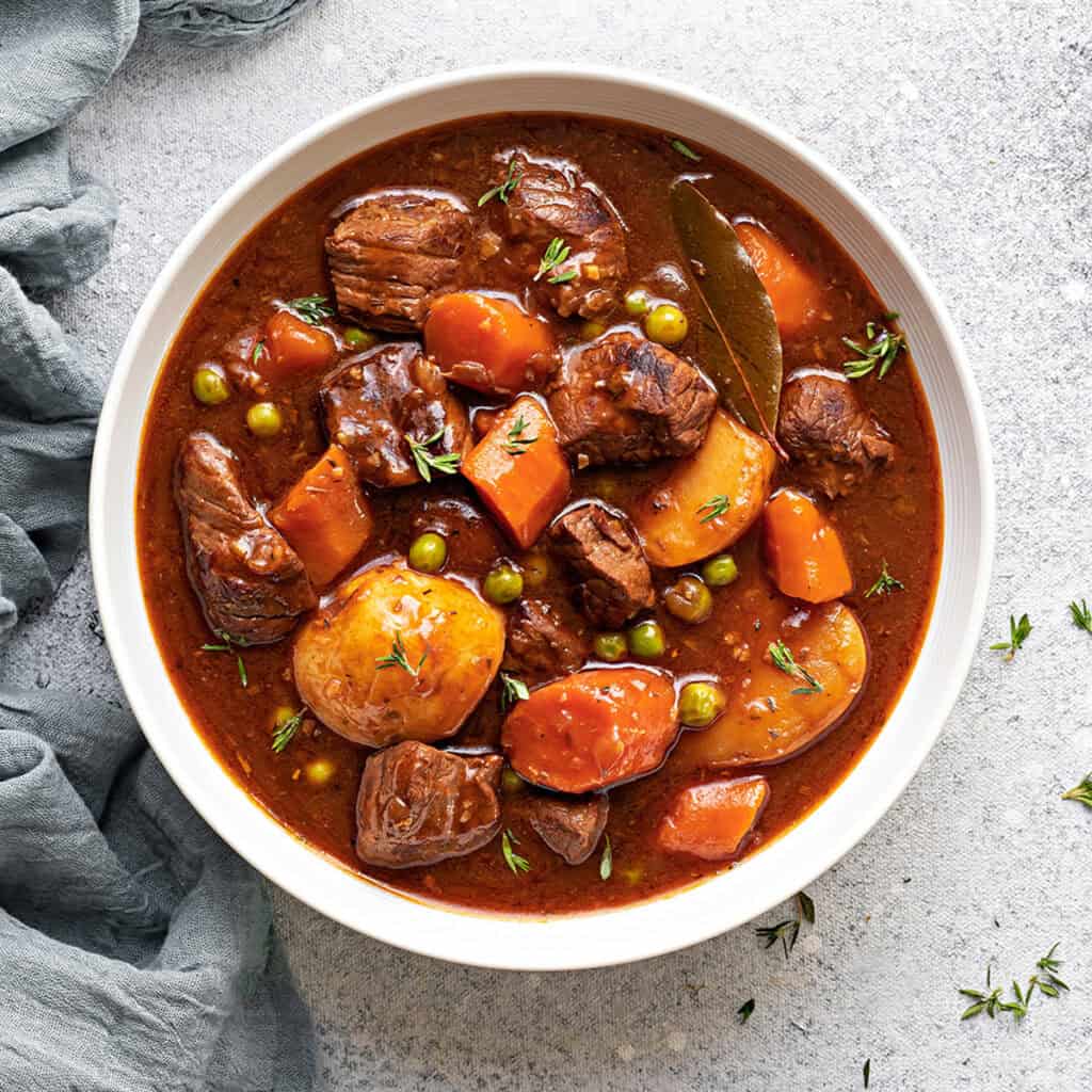 A white bowl of instant pot beef stew meat and gravy with potatoes, carrots and peas.