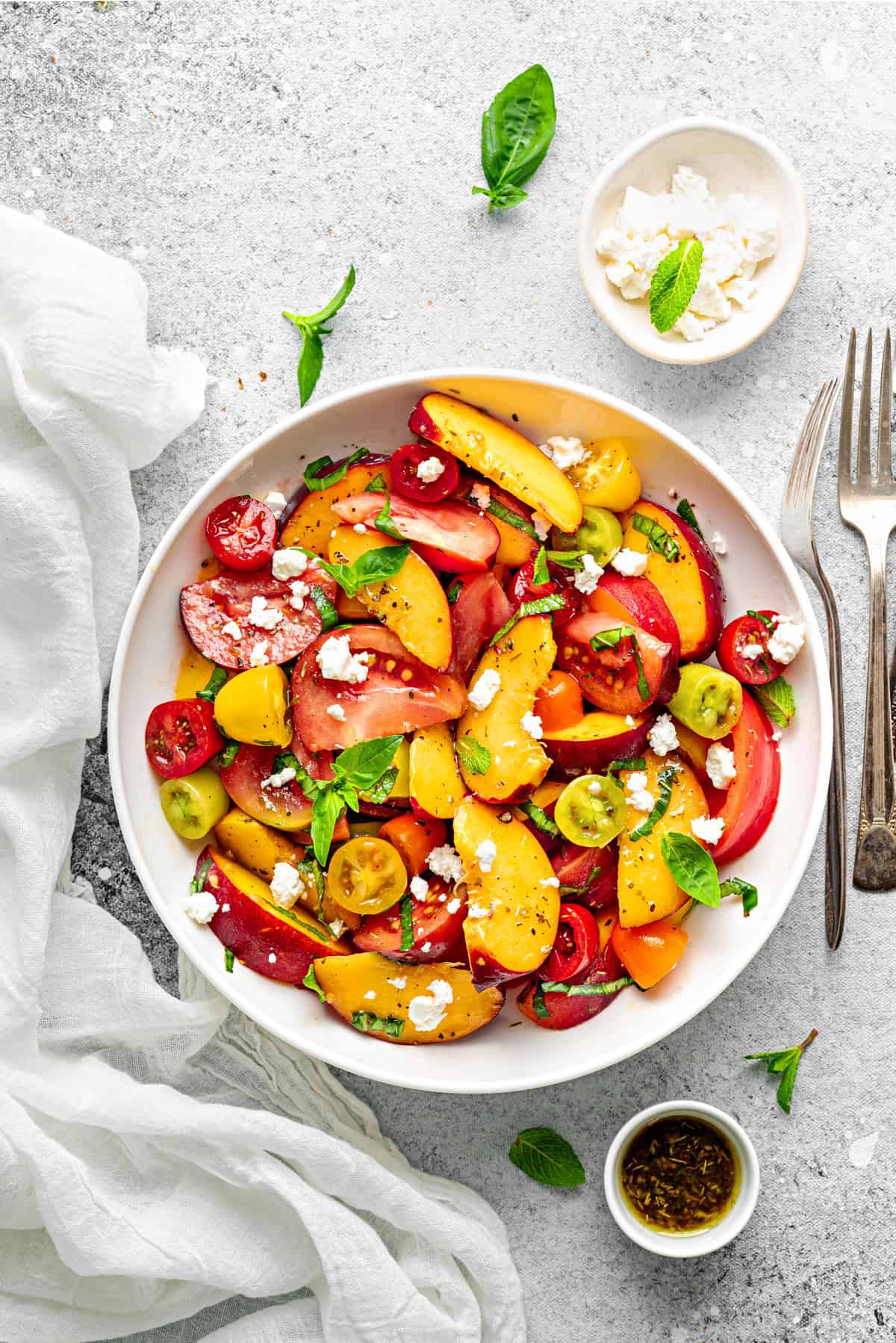 Top-down view of Peach Tomato Salad topped with feta, basil and mint in a white bowl for serving with a pair of fork on side.