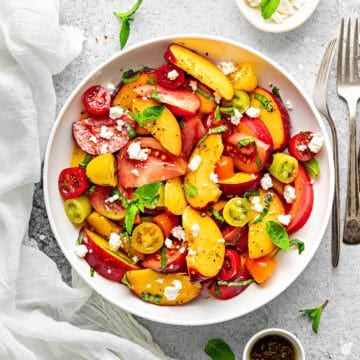 Peach Tomato Salad topped with feta, basil and mint in a white bowl for serving with a pair of fork on side.