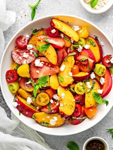 Peach Tomato Salad topped with feta, basil and mint in a white bowl for serving with a pair of fork on side.