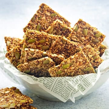 Close view of gluten-free mixed seed crackers stacked in a white ceramic bowl with parchment paper.