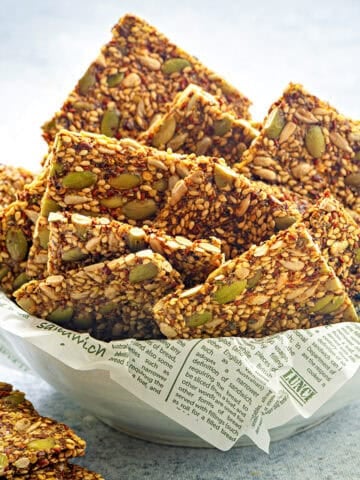 Close view of gluten-free mixed seed crackers stacked in a white ceramic bowl with parchment paper.
